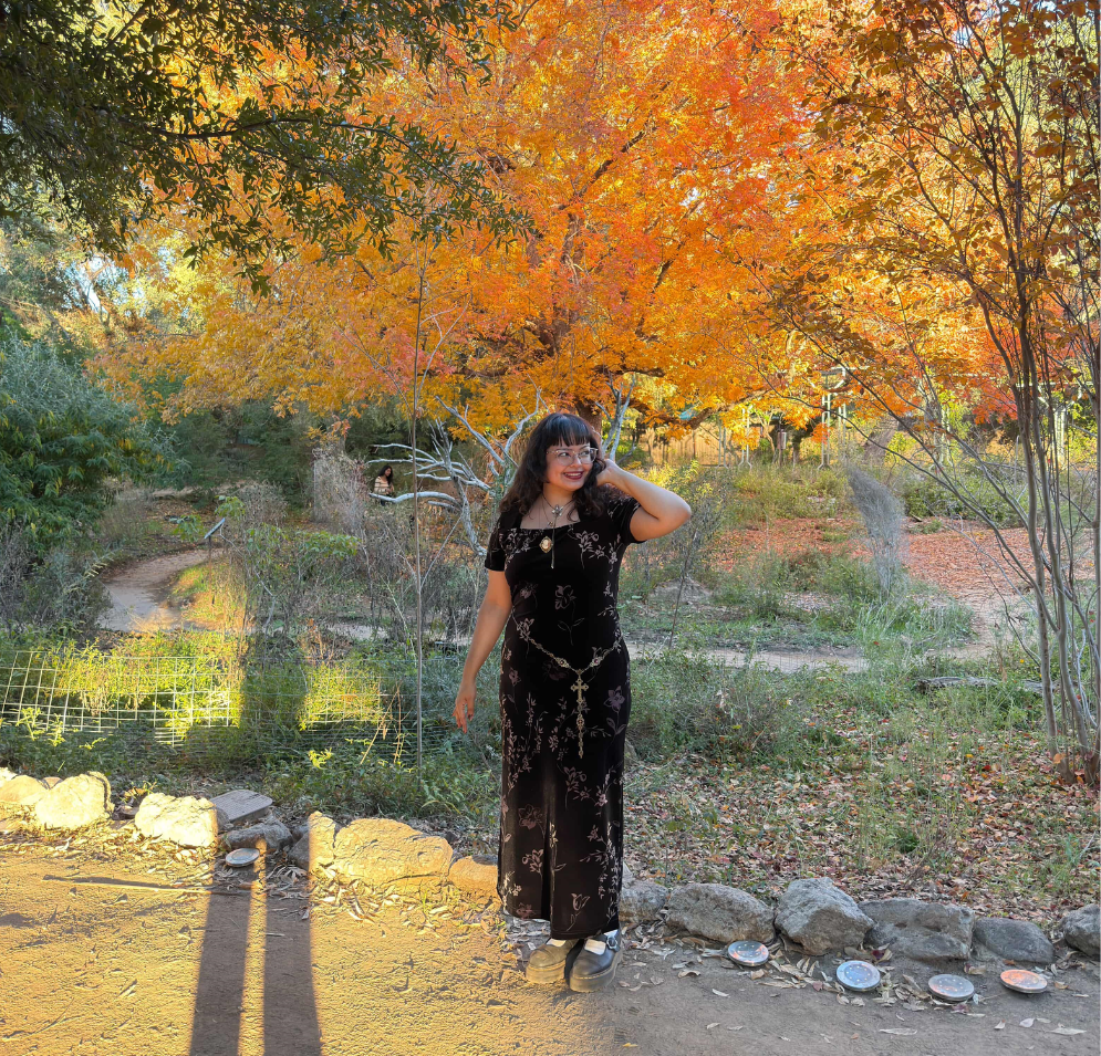 A woman with long dark hair is wearing a long dress in front of a tree with bright orange leaves.
