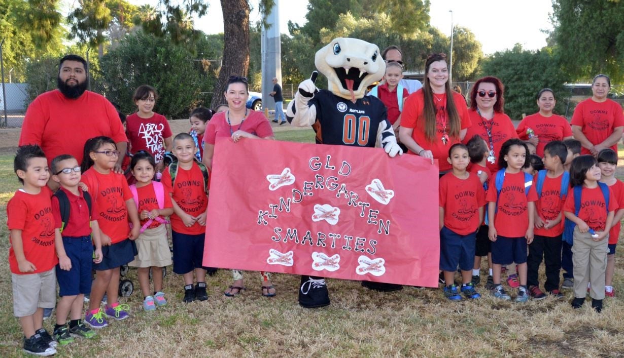 A group of elementary school children and adults gather outdoors on a grassy field, dressed in red shirts. They pose for a photo with the Phoenix Suns' gorilla mascot, who wears a white Suns jersey and squats in the foreground with arms outstretched. Some children hold black and orange Suns-themed bags. The background features trees, a speaker on a stand, and a crowd of parents and school staff. The setting is bright and sunny, with everyone smiling and engaged.
