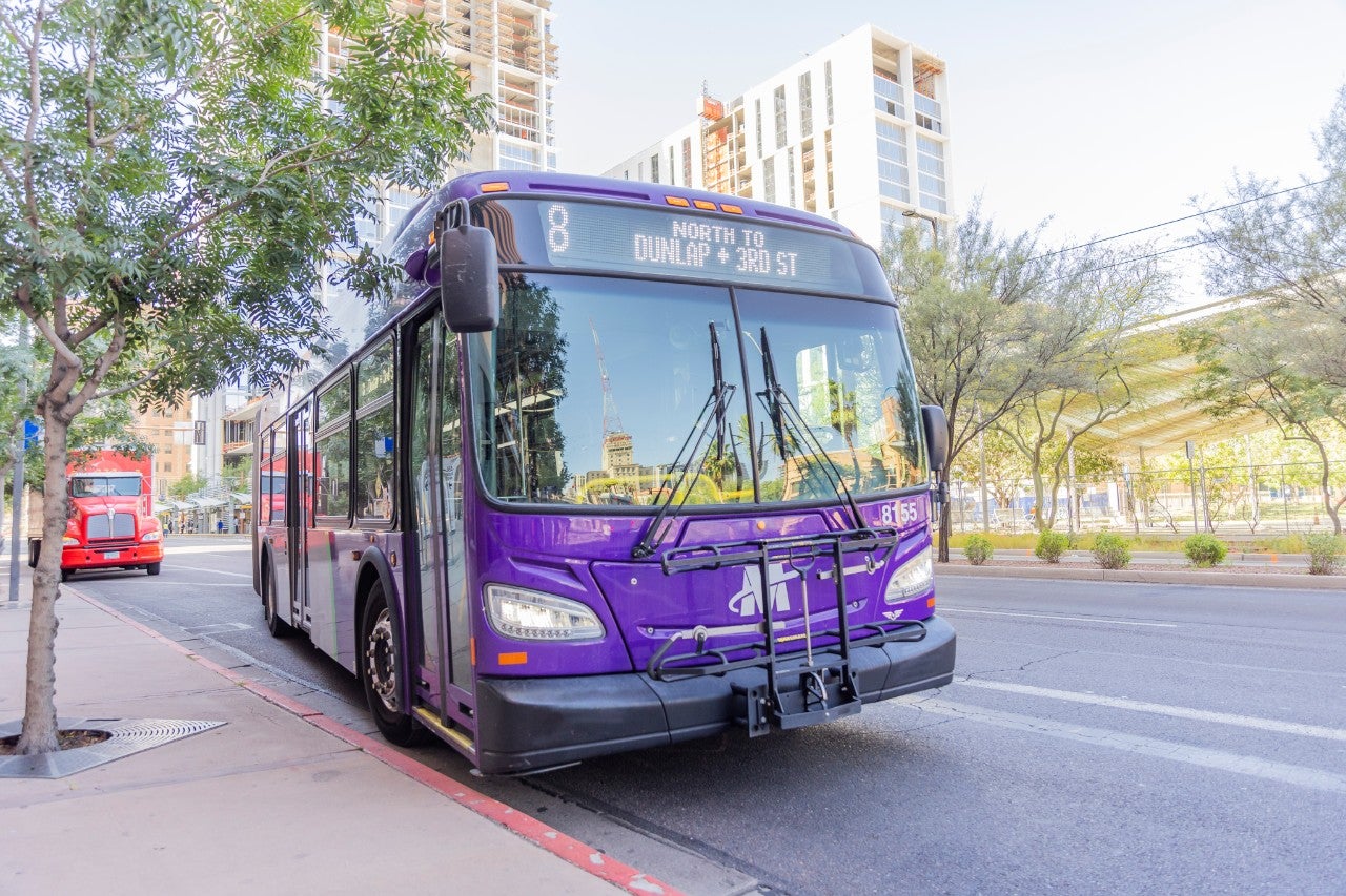 Purple bus pulling up next to a bus stop with tall buildings in the background.