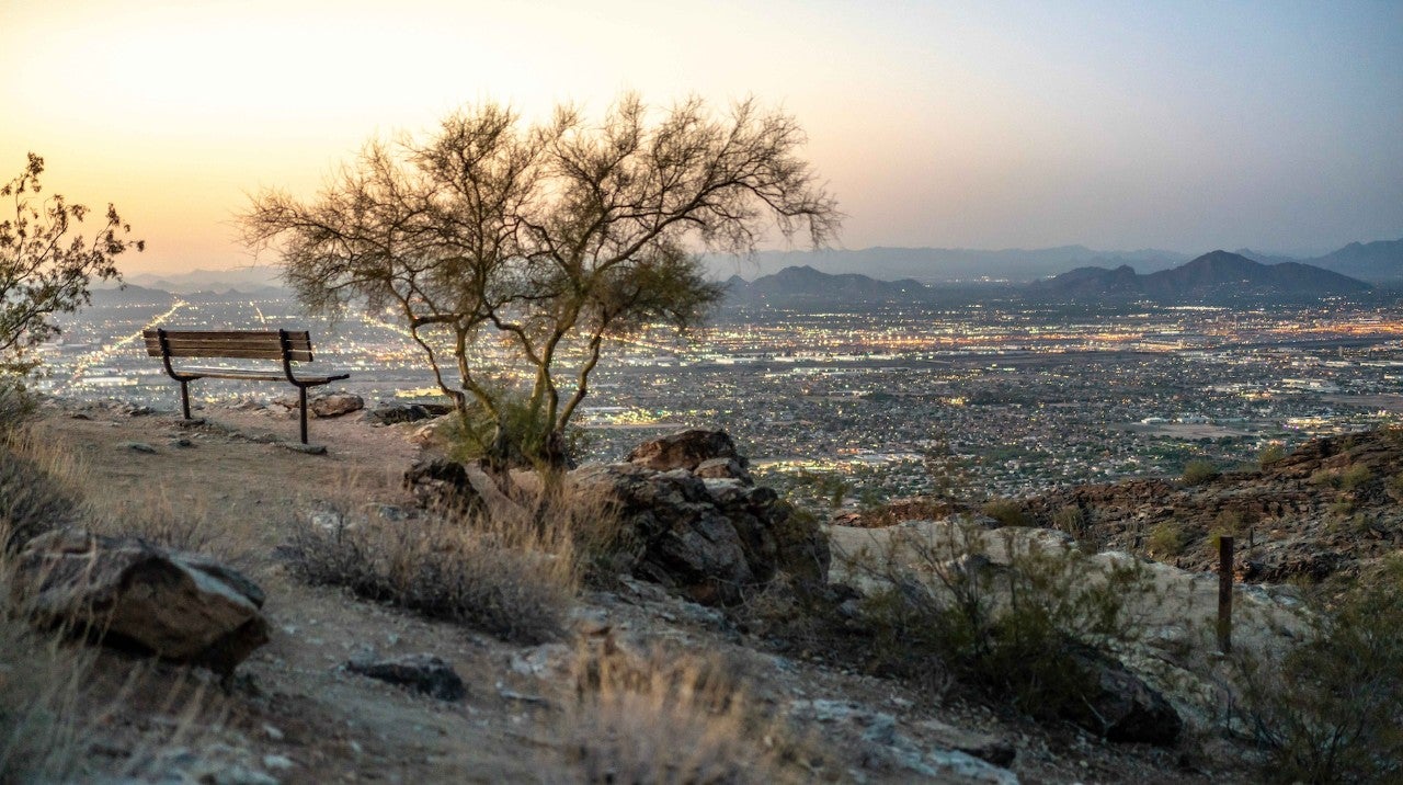 Photograph from South Mountain at dusk. It shows a a natural area with dirt, plants, and a bench overlooking city lights in the distance.