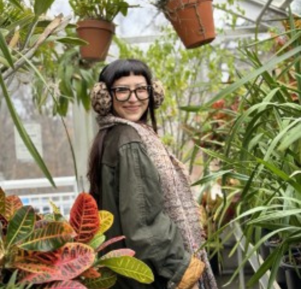 A woman with long dark hair, glasses, and earmuffs stands in  what appears to be a greenhouse, surrounded by various colorful, potted plants.
