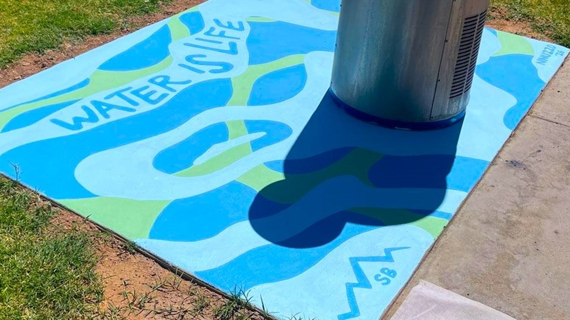 A patch of blue and green concrete surrounds the base of an outdoor drinking fountain. The artwork features the words "Water is Life" and several round, flowing shapes.