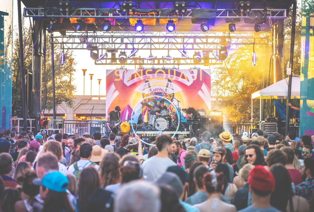 A crowd of people stand, chat, and listen to a band performing on a stage.