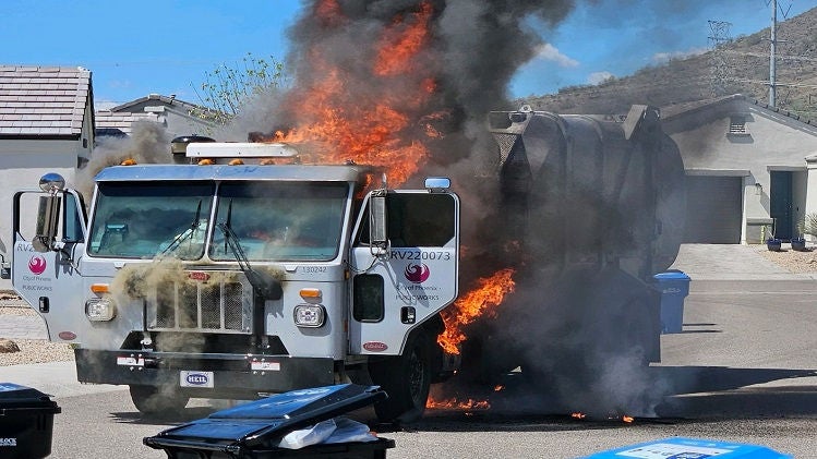 Flames and dark, heavy smoke engulf a Phoexni garbage truck in the street with trash and recycling bins in the foreground.