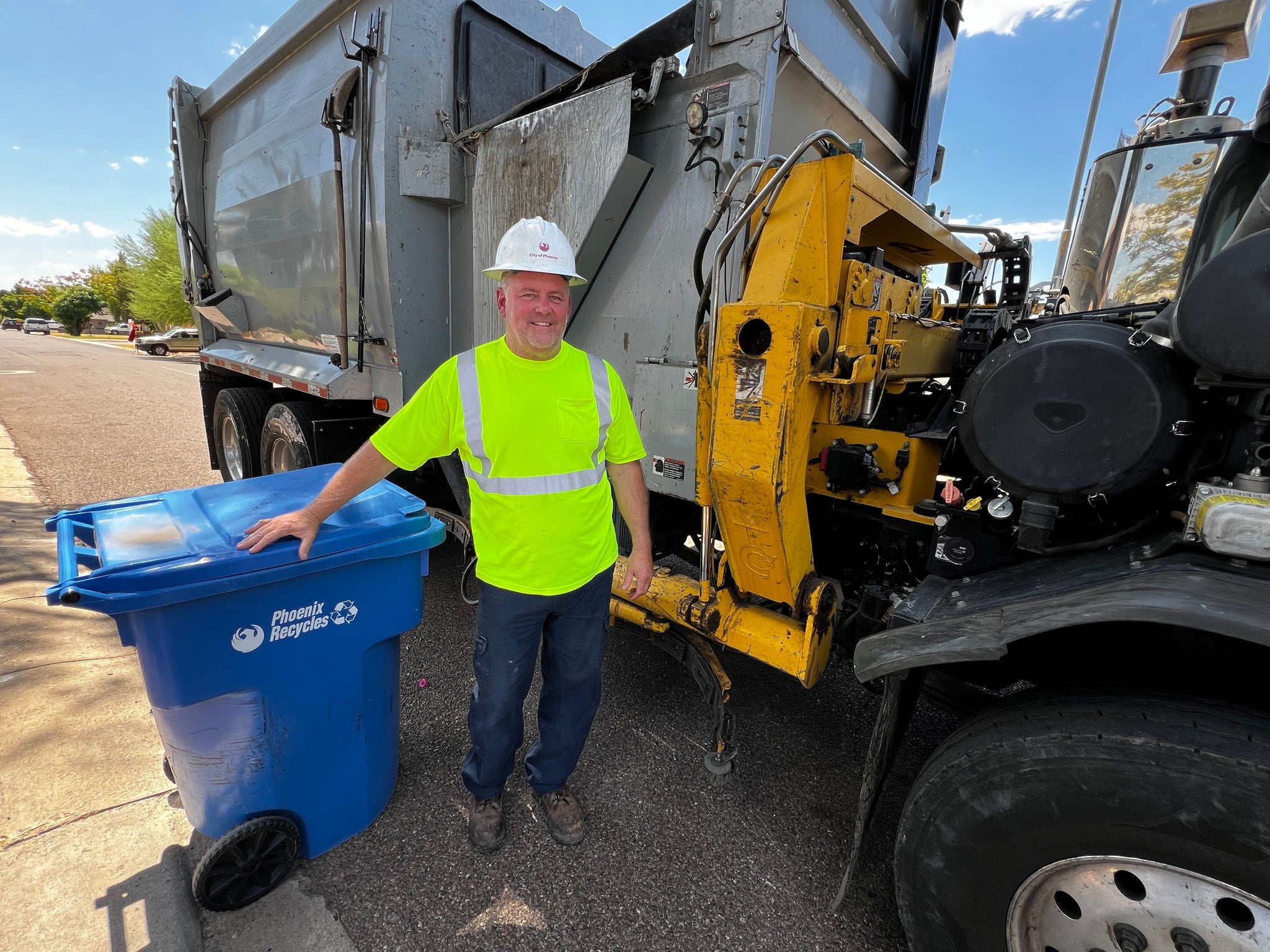 A solid waste equipment operator with a hard hat and reflective gear stands outside his truck next to a  blue recycle bin.
