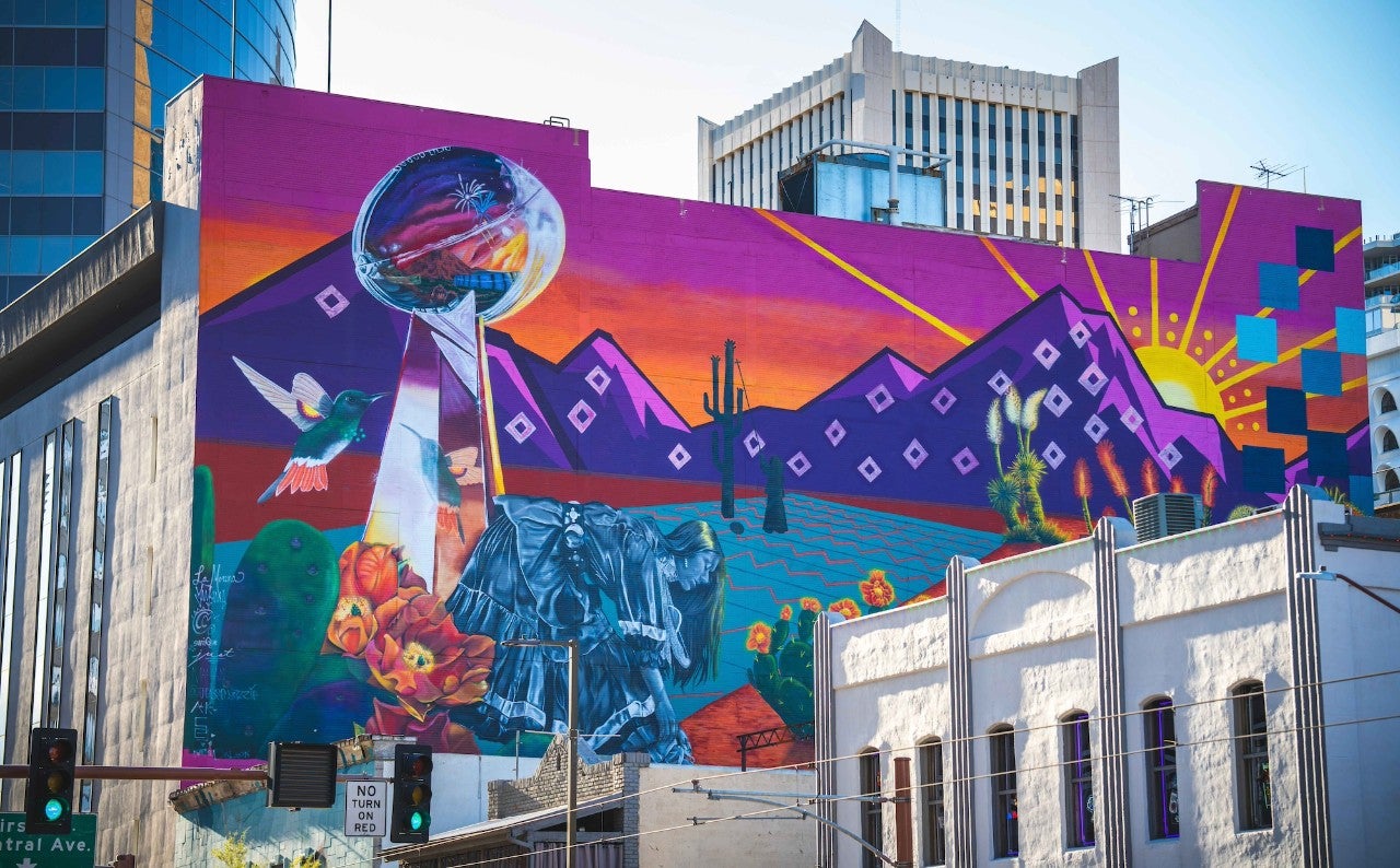 Mural on an urban building. The mural is saturated with pinks, purples, oranges, and blues. It depicts a mountain range, a body of water, a football trophy, cacti, and a Native American woman.