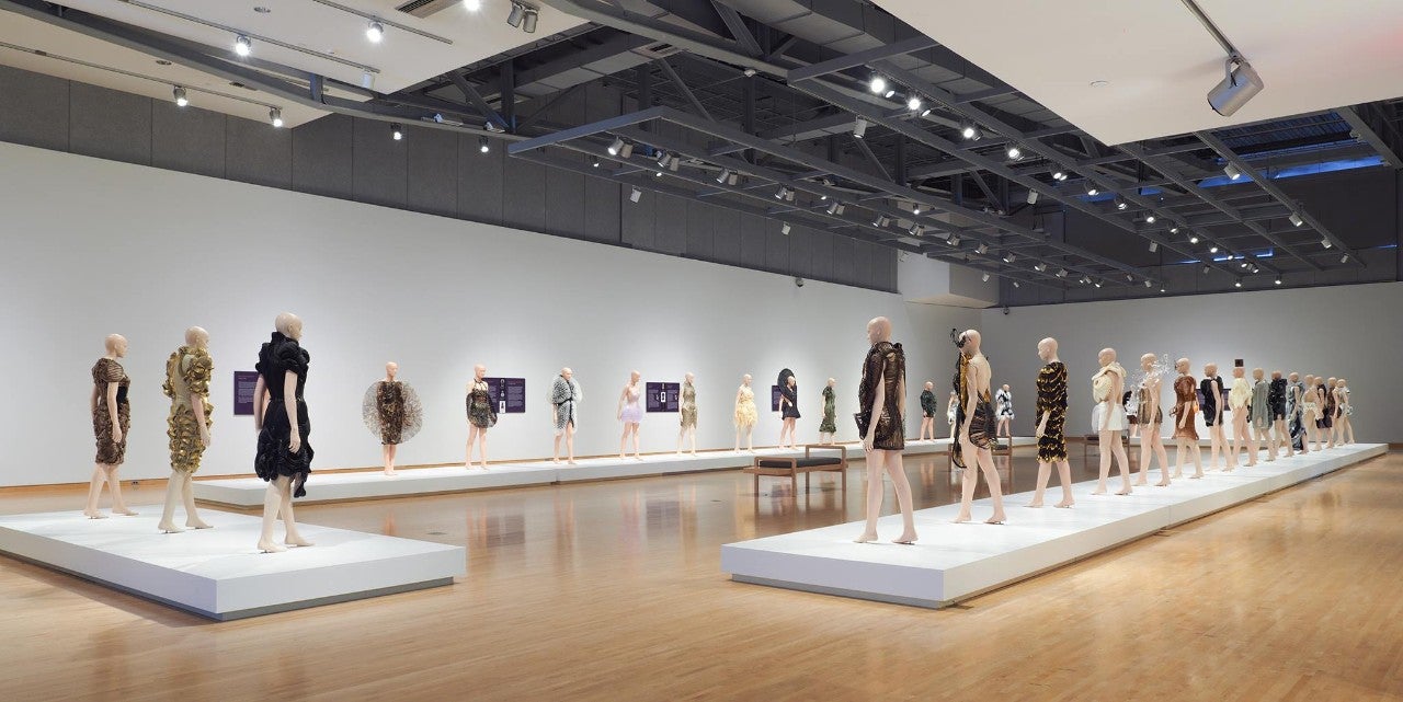 Multiple mannequins wearing fashion garb are arranged in a square in a museum exhibit.