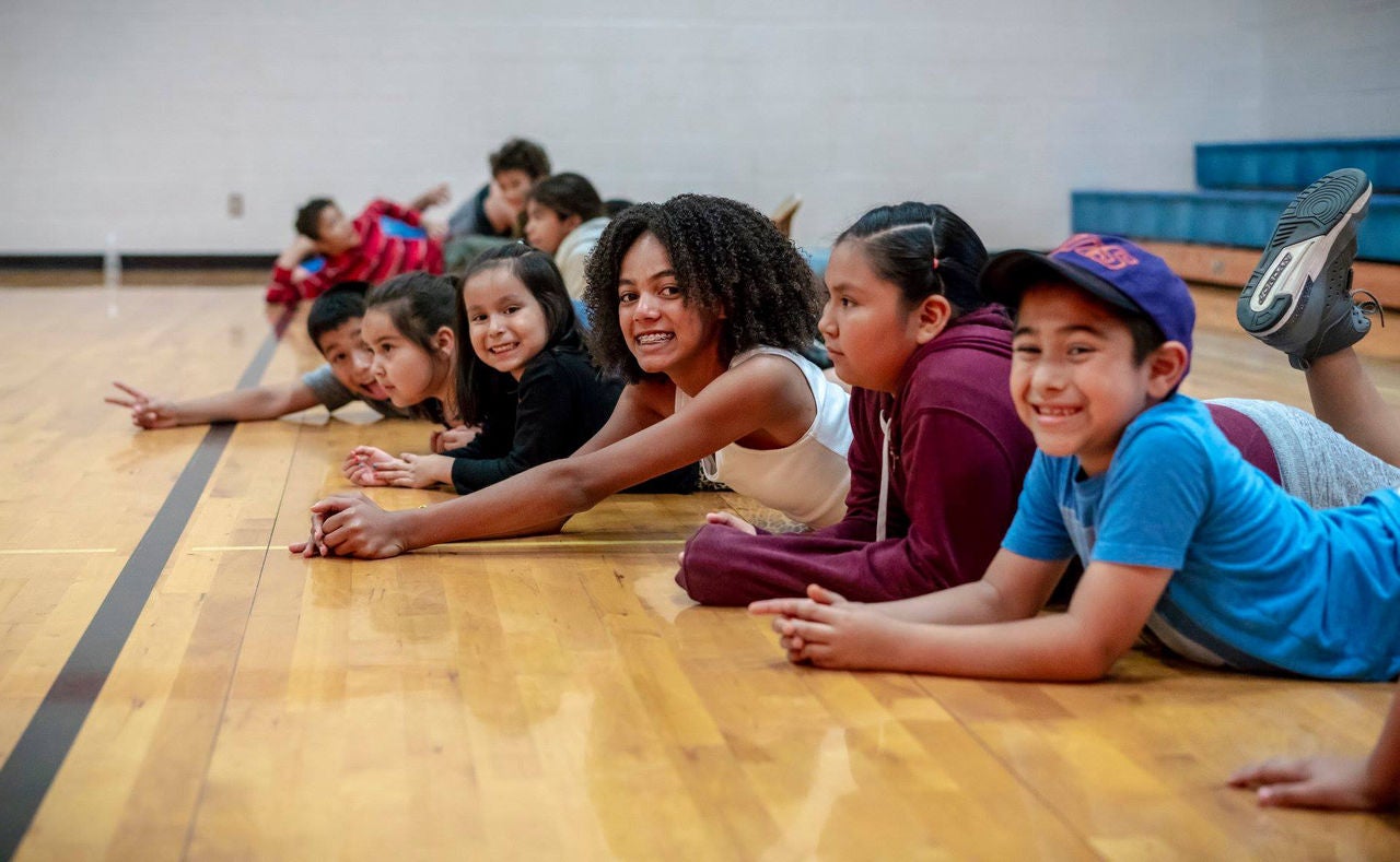 A group of children lay on the floor in a gym and smile at the camera.
