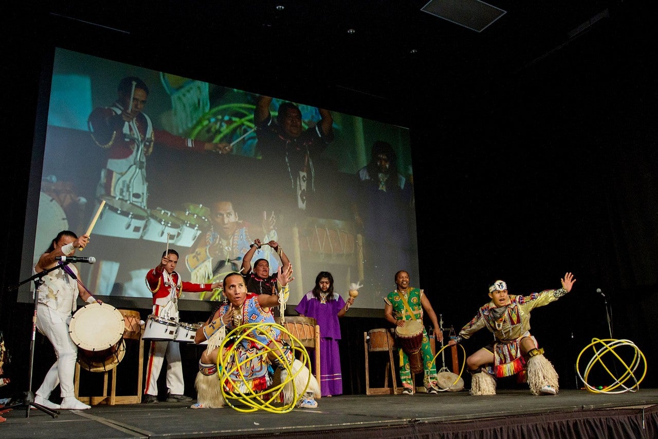 A group of American Indians wearing ceremonial outfits stand on a stage, play instruments, and dance.