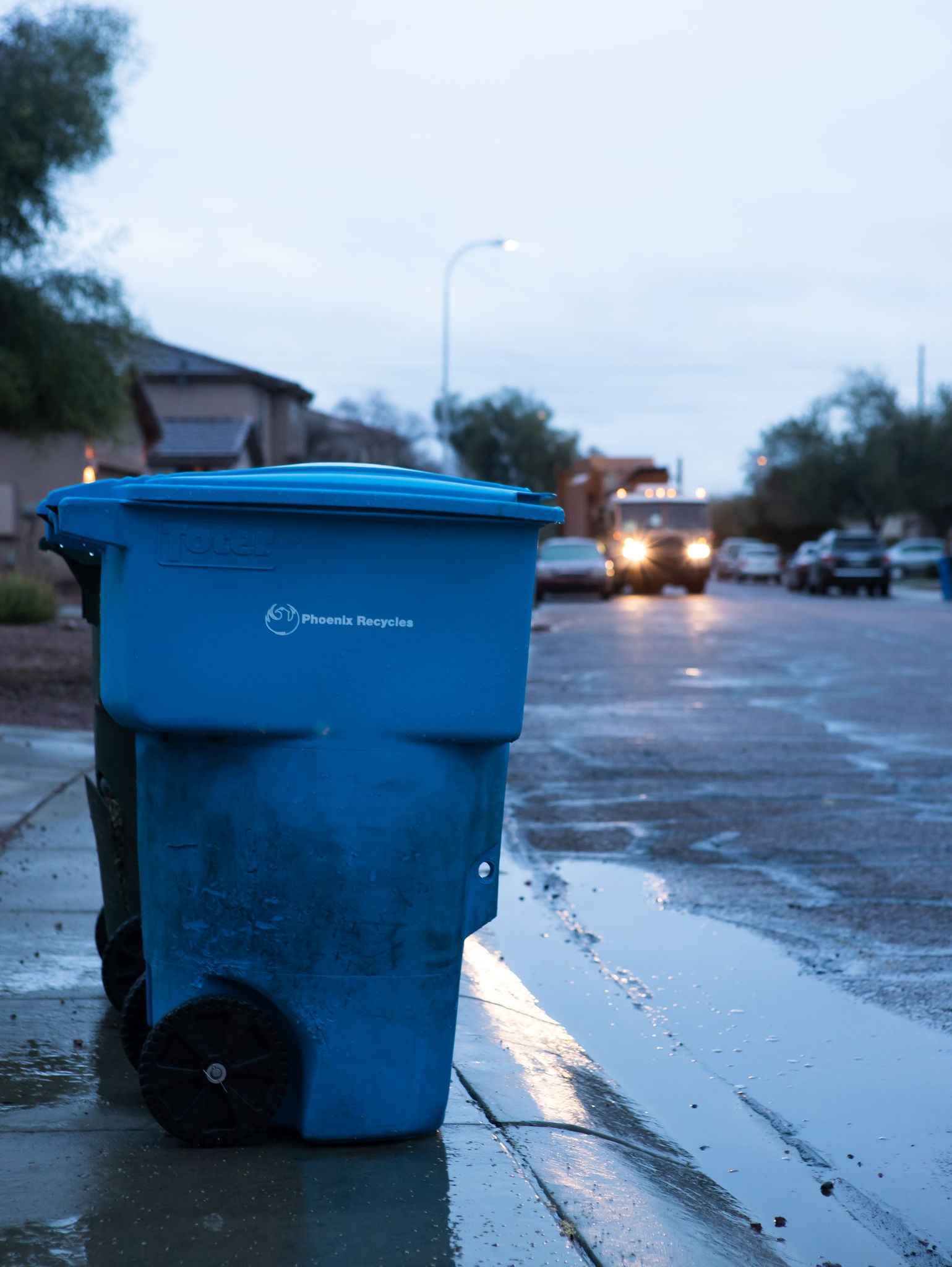 A recycle bin in front of a house on a rainy day. 