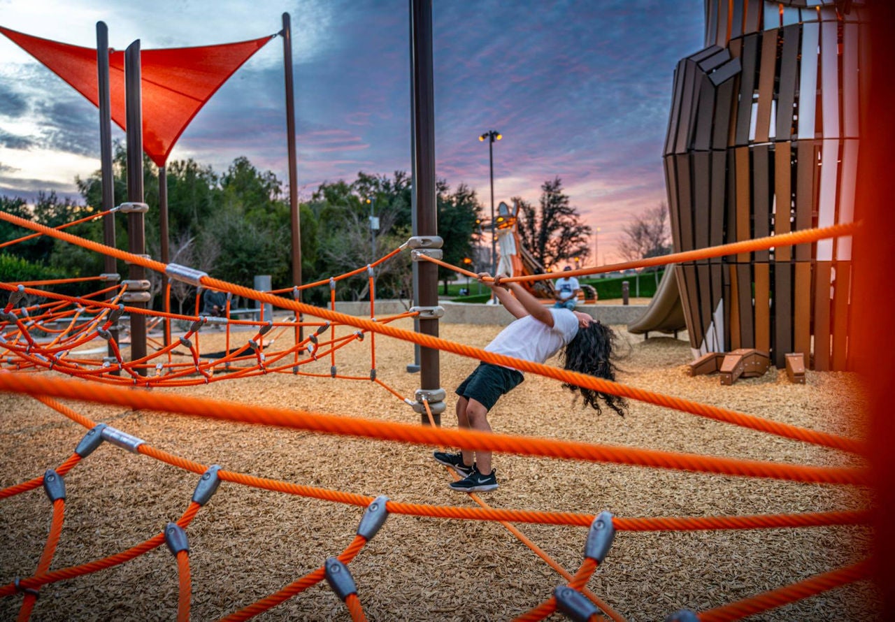 A child plays on an orange-roped jungle gym with more playground equipment in the background.