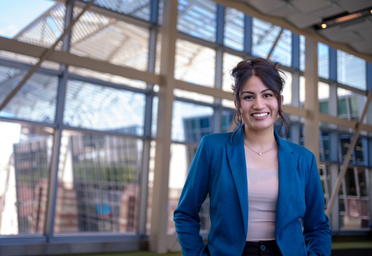 Woman in a blue blazer and brown shirt, blurred background of glass and steel and buildings beyond.