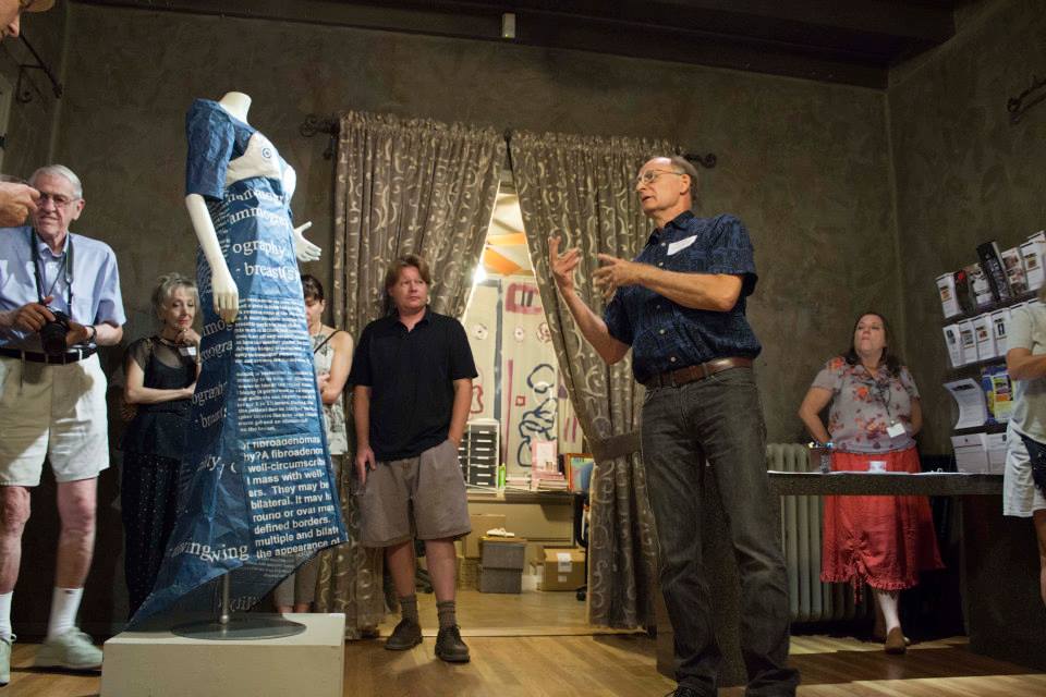 A man stands and spreads in front of a mannequin that is wearing a dress with text on it. Multiple people stand and listen to the man speak.