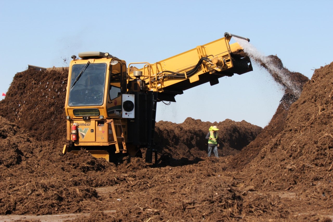 Large orange machine sprays dirt into a pile. A person who is half the size of the machine and is wearing a neon yellow vest looks on.