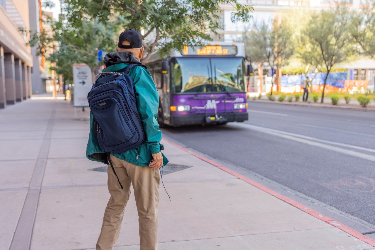 Man in a green jacket and black cap waits by a bus stop as a Valley Metro bus pulls into the stop. 