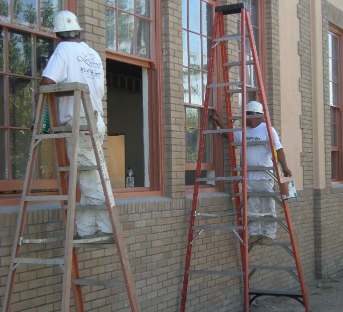 Two men stand on ladders and work on a building's windows.
