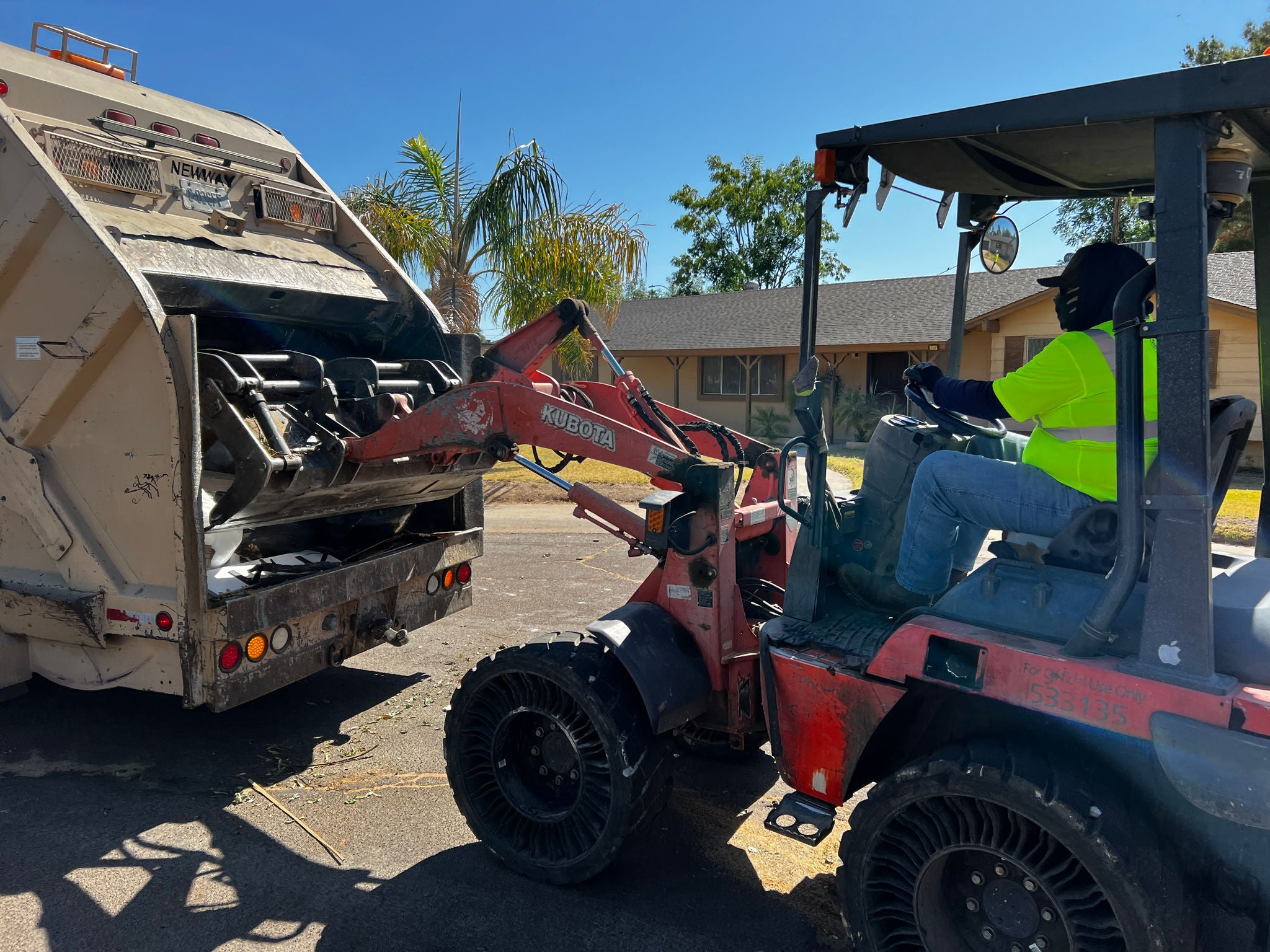 A solid waste equipment operator in bright yellow reflective gear uses a tractor to place bulk trash items into a rear-loader garbage truck.