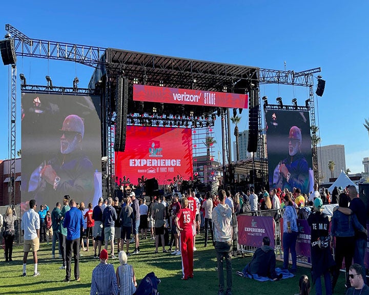 Outdoor event with a stage stage featuring a performance, large video screens, and an audience on the grass.