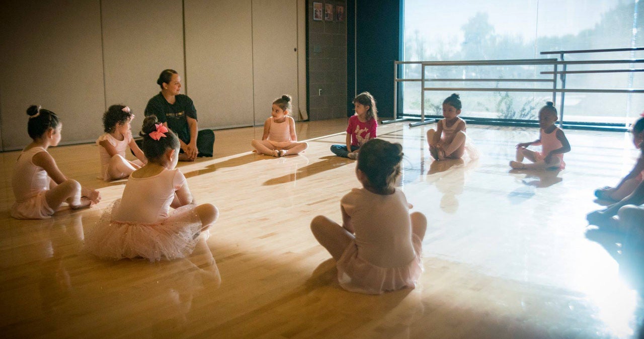 A group of young girls in ballerina girls sit in a semi-circle on the ground next to an instructor.
