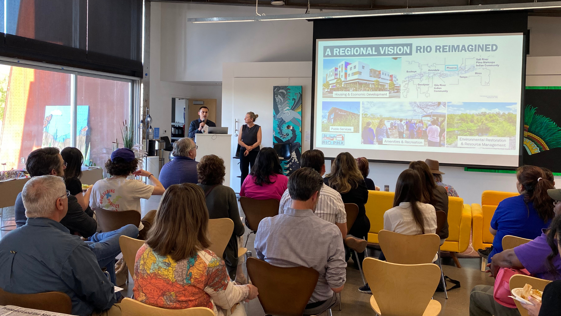 A presentation taking place in a modern room with a projector screen displaying 'A REGIONAL VISION RIO REIMAGINED' and several images. Two presenters stand near the screen, while an audience sits facing them.