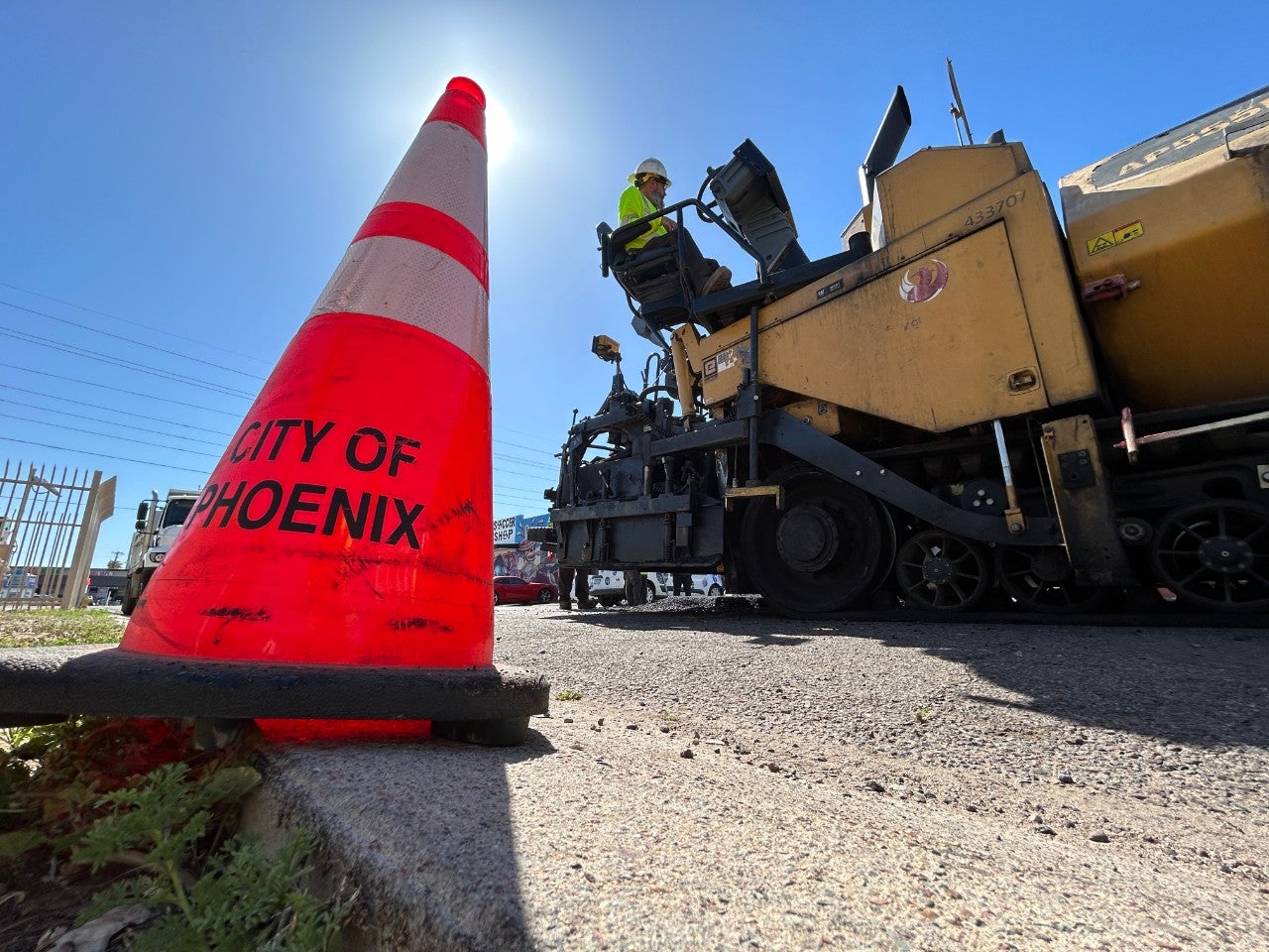 Low-to-the-ground photo of a bright orange City of Phoenix traffic cone, with construction workers on a large vehicle in the background.