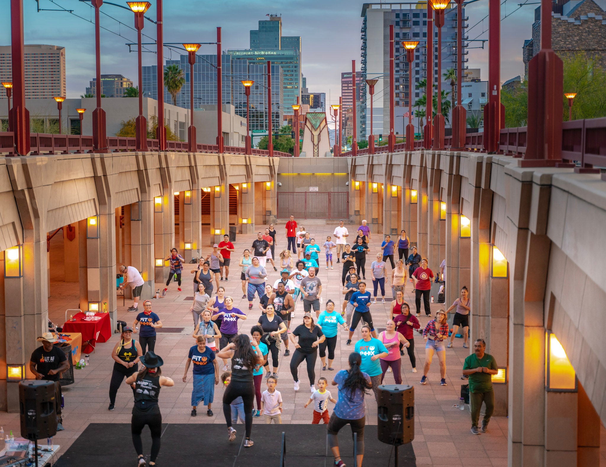 A crowd of people face a stage and follow instructions for a group exercise class. They are standing in a common area set below the road, and the Phoenix skyline is visible in the distance.