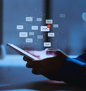 A close-up shows hands holding a cellphone. Above the device, several digital envelope icons float in the air, representing a large number of incoming emails. The image suggests high email activity, possibly related to spam or phishing scams.