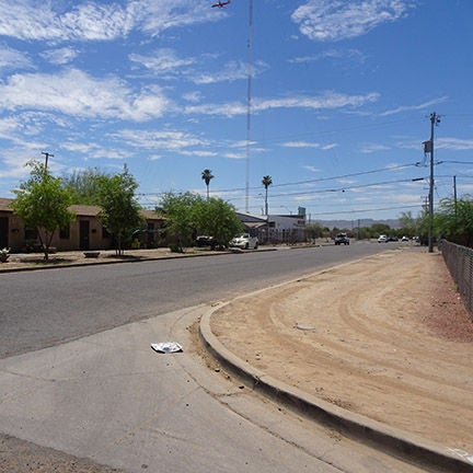 A corner at an intersection on a road with no visible lane markings. Treadmarks in the dirt beside the road indicate people have been using it as a walking or biking path.