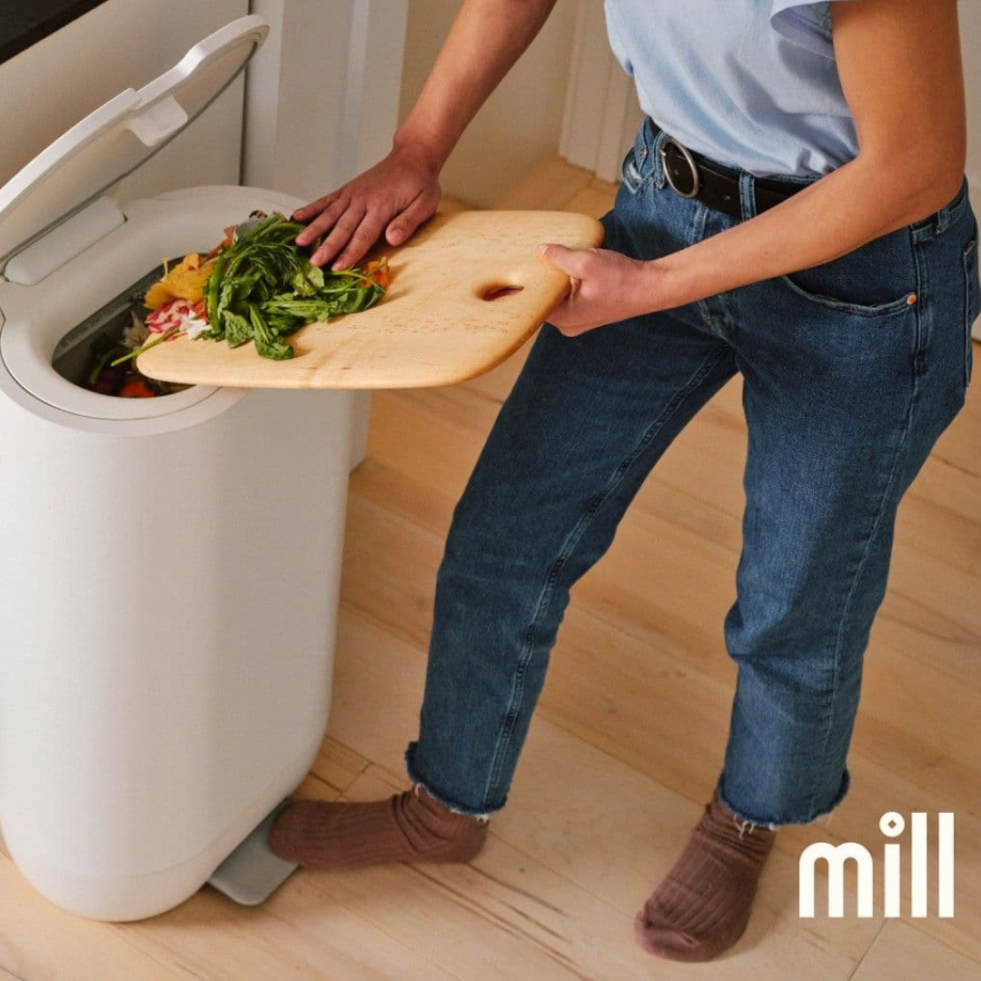 A woman slides food scraps from a cutting board into a kitchen food recycler -- a white bin about the size of a standard kitchen trash can.