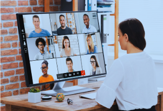 A woman sits at a desk wearing a white blouse with three-quarter sleeves. Her dark hair is swept to the right side of her head, and she is facing a computer monitor with her back to the viewer. The screen displays a video conference with nine people arranged in three rows—five men and four women. The image represents a virtual meeting.