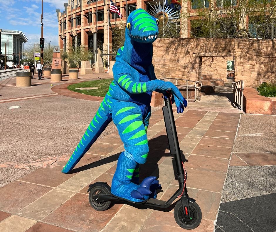 Zero, a blue and green dinosaur mascot, stands on an electric scooter in front of Phoenix City Hall.