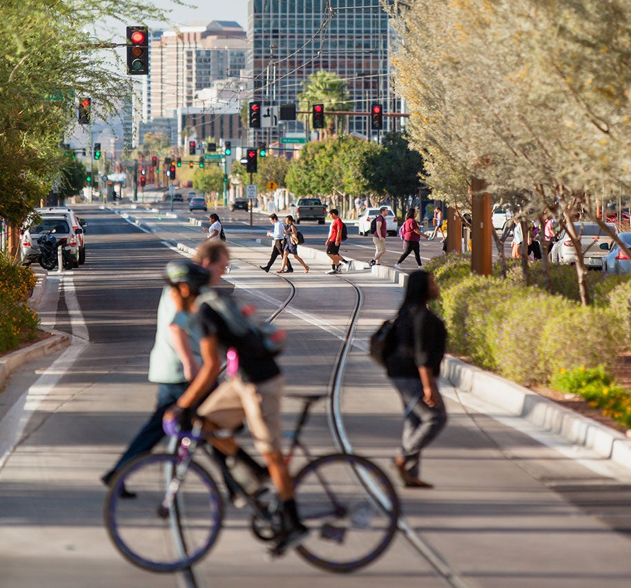 Pedestrians and cyclists navigate a busy urban street with traffic lights, crosswalks, and rail tracks, showcasing a vibrant cityscape in motion.