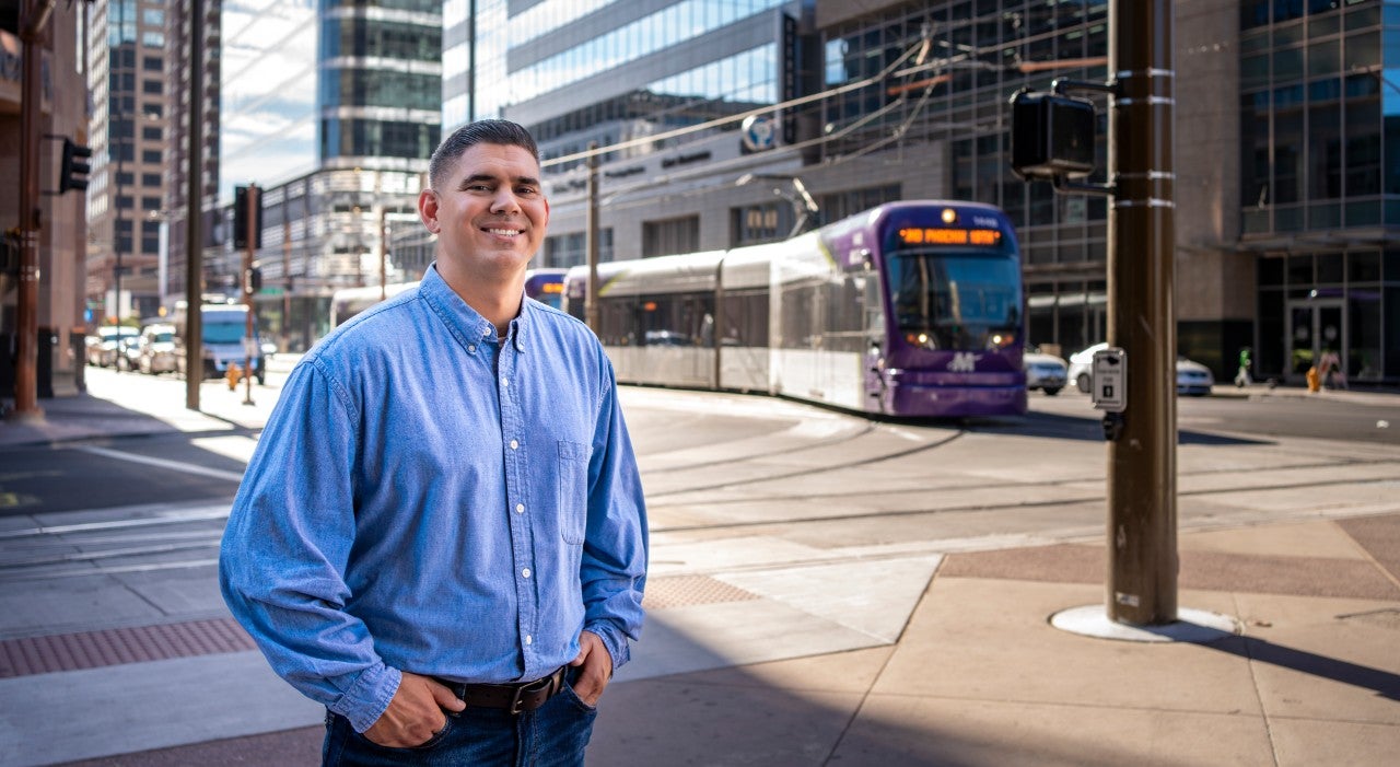 A man in a blue dress shirts smiles at the camera in front of a busy Phoenix street.