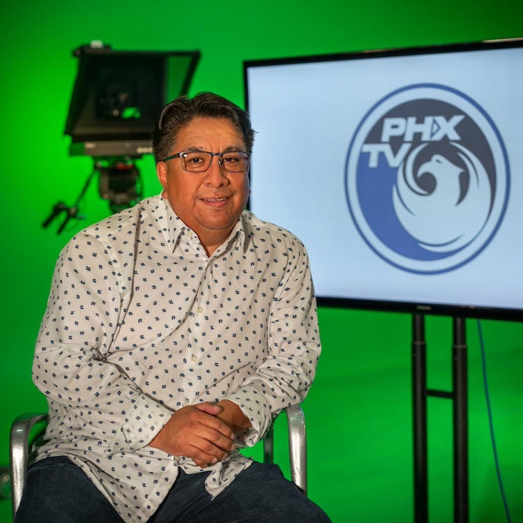 Felipe Peña sits in front of a green screen and television studio equipment, including a large television that shows the PHXTV logo.