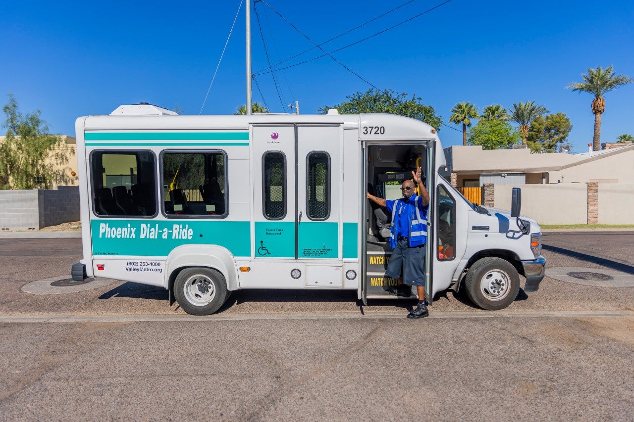 A Dial-a-Ride bus operator waving as he stands in front of a white and blue Dial-a-Ride bus. 