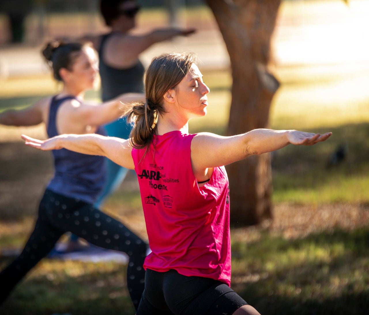 Three individuals in a yoga pose with their arms straight out during an outdoor class.