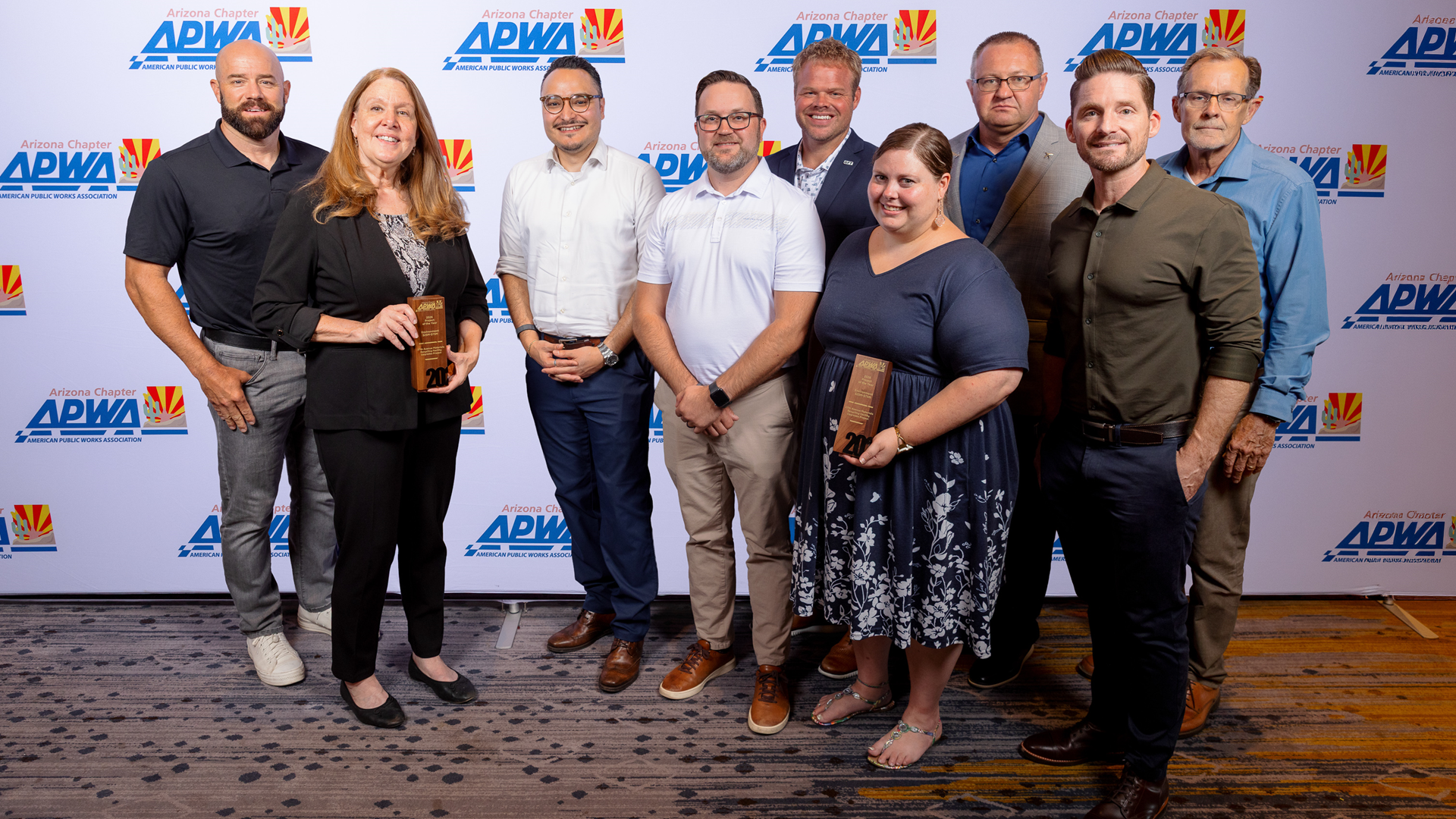A group of 9 adults in professional attire pose indoors with a trophy in front of a photo backdrop that features the repeated logo of the American Public Works Association..