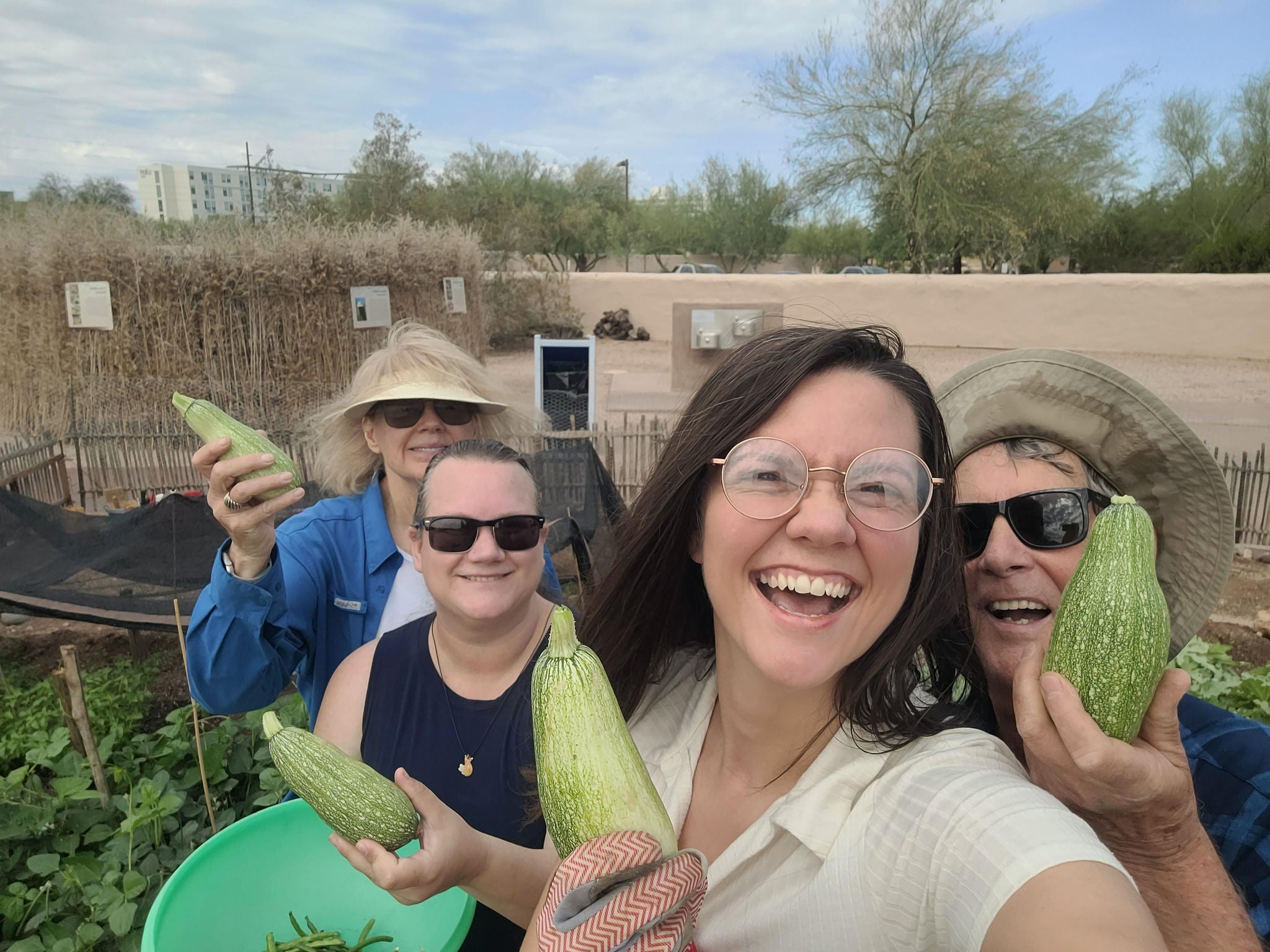Group photo of four garden volunteers during an autumn harvest. Each one smiling, wearing sunglasses, and holding zucchinis in one hand. Greenery from the rest of the crops and the museum's perimeter wall in the background.