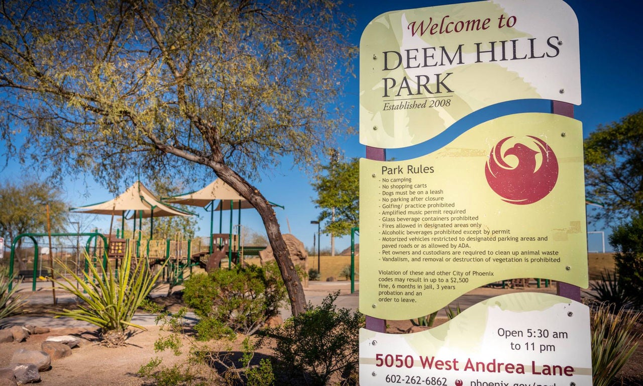 Welcome sign for Deem Hills Park with greenery and playground equipment in the background.