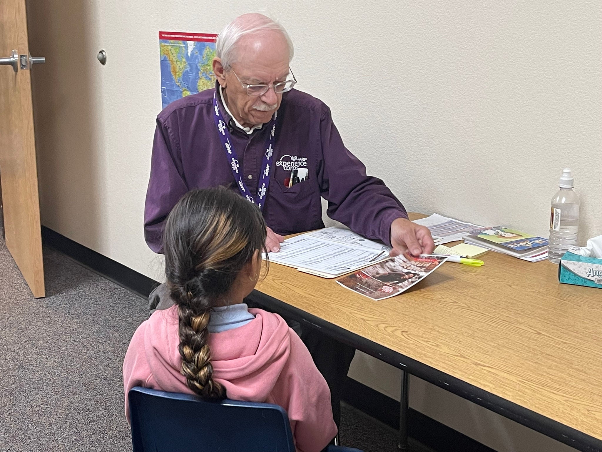 A young student and an adult sit together at a rectangular table in either a school or office setting. The adult has a colorful flyer and other paperwork on the desk in front of him.