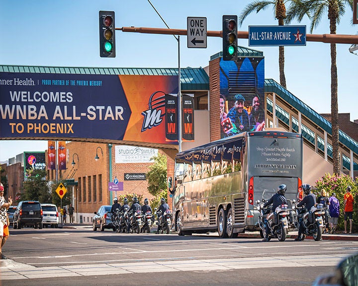 Street scene in Phoenix with a banner welcoming the WNBA All-Star event, featuring a bus, police motorcade, and visible signage for 'All-Star Avenue'.