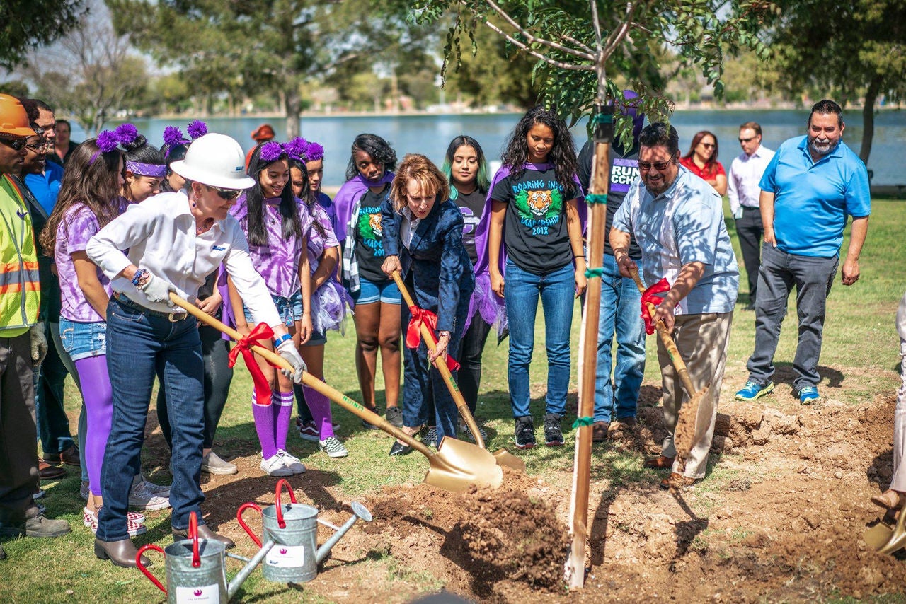 Three individuals plant a tree while a group of onlookers watch.