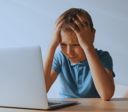 A young boy sits at a table facing a silver laptop. He is wearing a blue casual top, with his arms resting on the table and his hands pressed against his head. He looks scared and upset and crying. He is alone and looks terrified. 