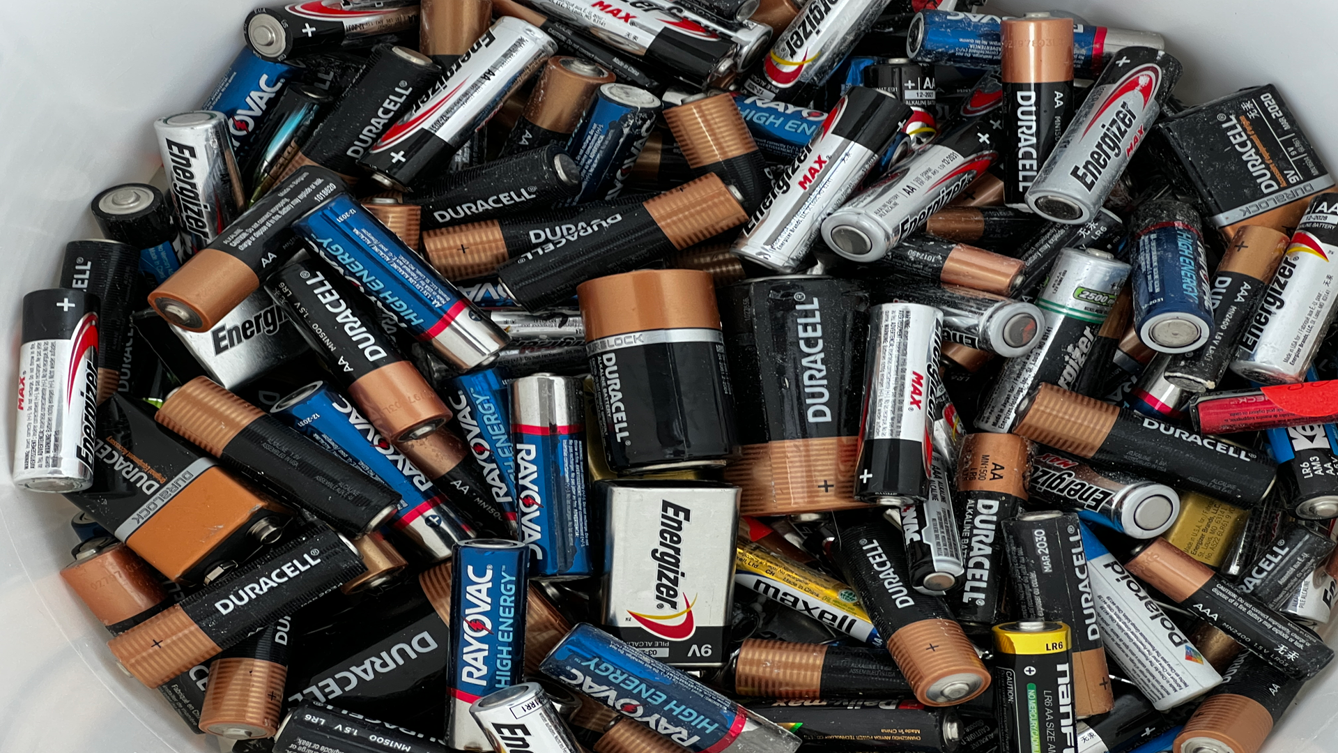 A white bucket filled with various brands and sizes of alkaline batteries.