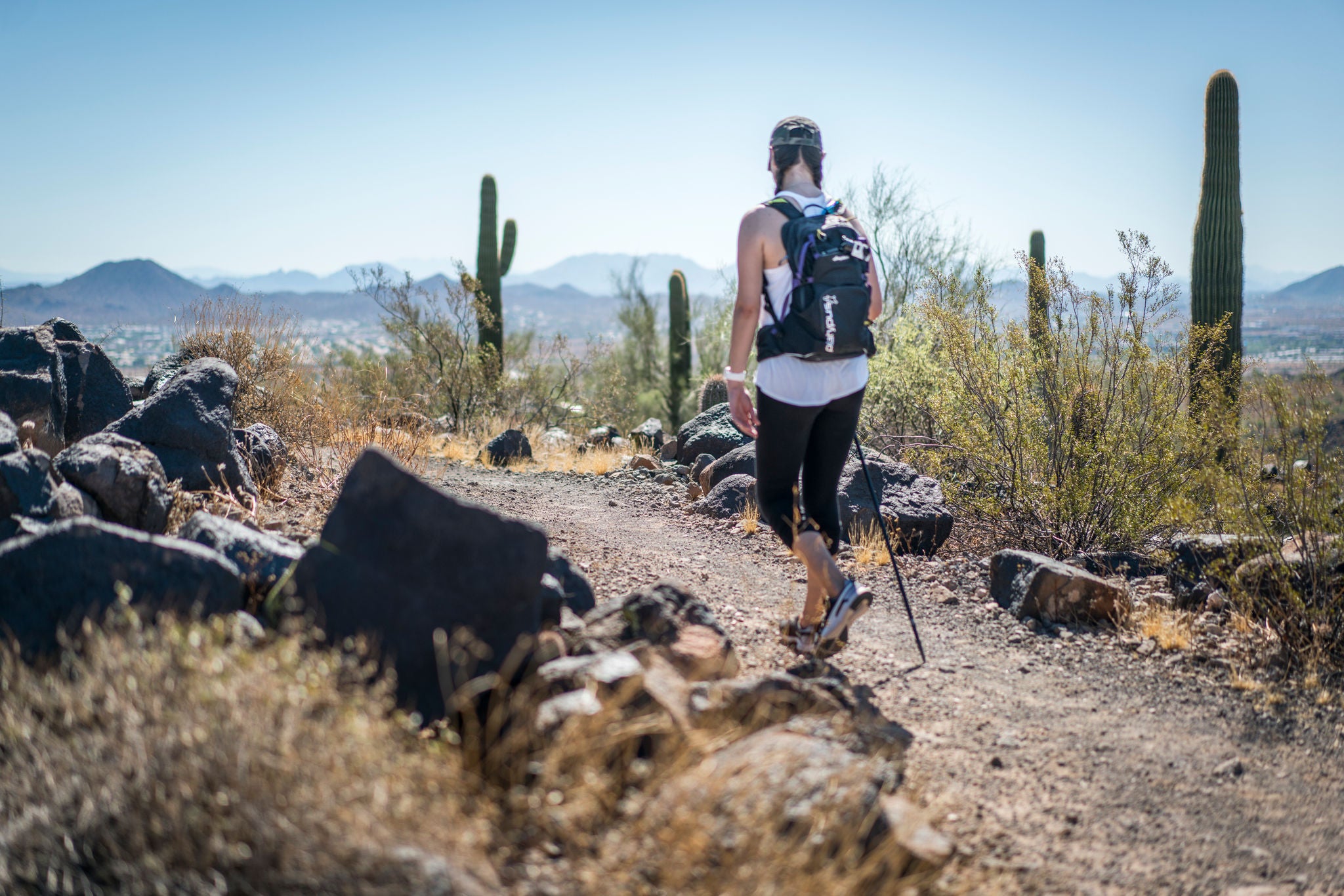 A hiker walks in Deem Hills Recreation Area with cacti, scrub, rocks, and  mountains in the distance.
