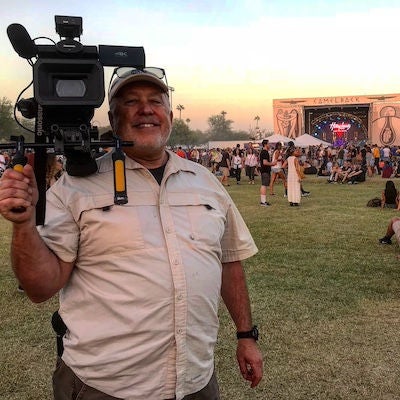 Matt Barcellos stands and smiles in front of an outdoor festival area. He is holding a large videocamera on his shoulder.
