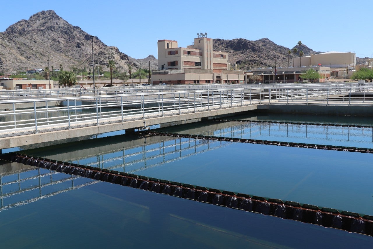 Water in a City of Phoenix Water Services Department water treatment plant with buildings, equipment, walkways for operators, mountains, trees, and blue sky in the background.