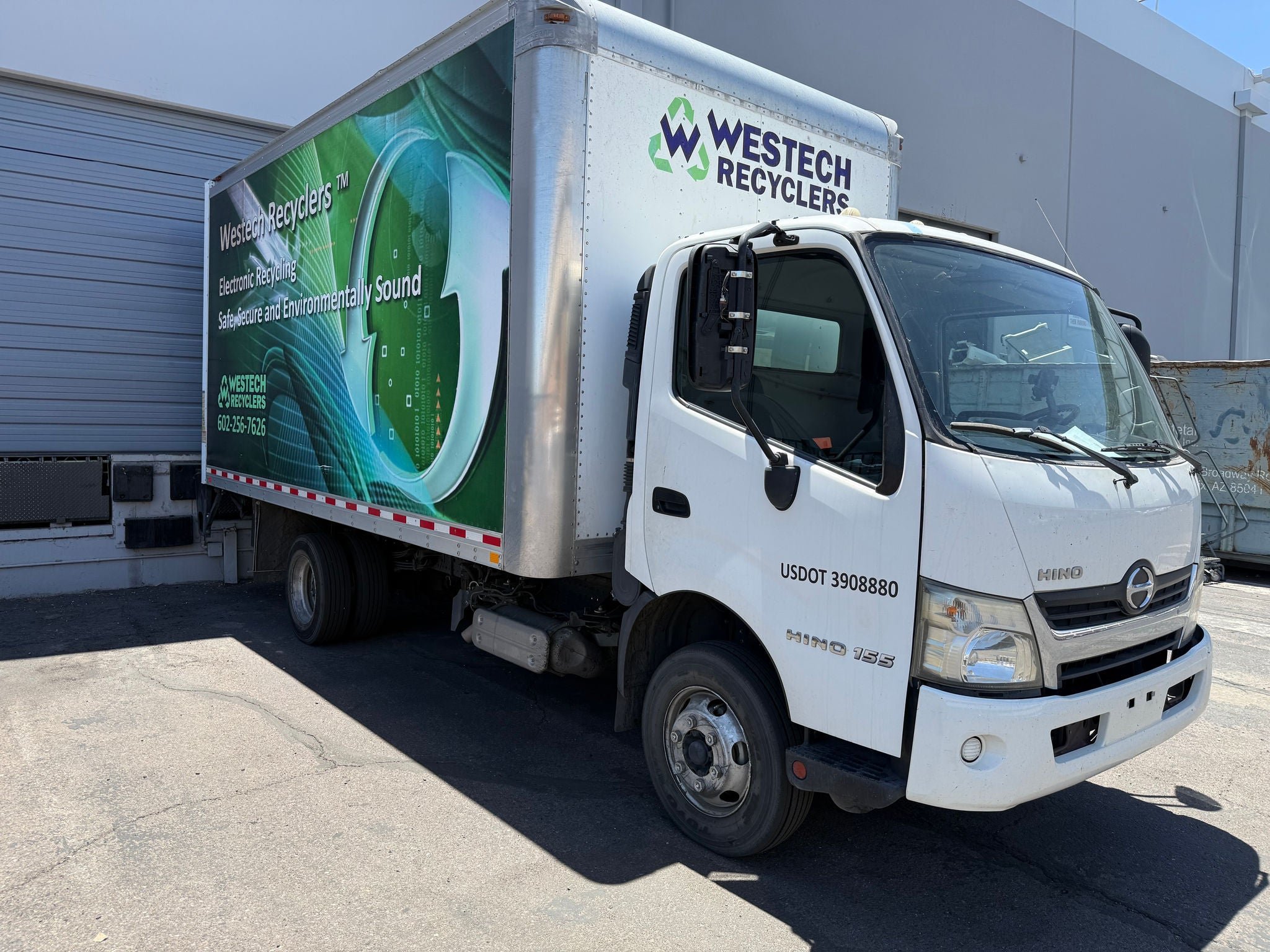 A white box truck with blue lettering that reads 'Westech Recyclers' is backed up to a loading dock on a sunny day.