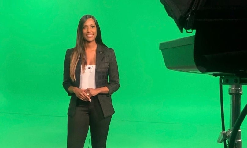 Davronja Dossie stands in front of a green screen and smiles as she's being recorded during a broadcast.
