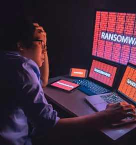 A man sits at a table in a dark room, surrounded by devices, including a cellphone, tablet, laptops, and a desktop computer. The only light comes from the screens, all showing the word "RANSOMWARE" in bold white letters. The man looks distressed, suggesting he is experiencing a cyberattack or data loss.